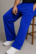 Load image into Gallery viewer, A person wearing bright royal blue sweatpants with white sneakers, seated on a wooden stool against a plain gray studio background.
