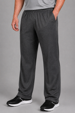 Load image into Gallery viewer, A person wearing dark gray heather lounge pants with an elastic waistband and side pockets, paired with black and white sneakers, shown from the waist down against a solid gray studio background.