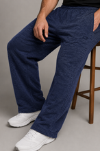 Load image into Gallery viewer, A person seated on a wooden stool wearing loose, wide-leg navy pants and white sneakers against a plain gray studio background.