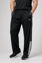 Load image into Gallery viewer, Black athletic track pants with white side stripes and a white logo, worn by a model in black sneakers against a light gray studio background.

