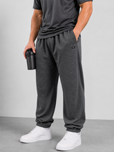 Load image into Gallery viewer, A person wearing dark gray heather jogger sweatpants with elastic cuffs, paired with a dark gray t-shirt and white sneakers, holding a black shaker bottle against a light gray studio background.
