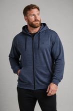 Load image into Gallery viewer, A man wearing a heathered navy blue zip-up hoodie with black drawstrings and a black front zipper, paired with black pants, photographed against a solid gray studio background.

