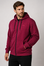 Load image into Gallery viewer, A man wearing a heathered burgundy zip-up hoodie with black drawstrings and a black front zipper, paired with black pants, photographed against a neutral gray studio background.
