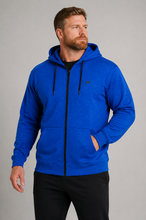 Load image into Gallery viewer, A man wearing a bright blue zip-up hoodie with black drawstrings and a black front zipper, paired with black pants, photographed against a neutral gray studio background.
