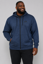 Load image into Gallery viewer, A man wearing a heathered navy blue zip-up hoodie with a small black chest logo, paired with black sweatpants, photographed against a solid gray studio background.

