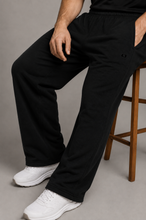 Load image into Gallery viewer, A person seated on a wooden stool wearing loose black sweatpants and white sneakers against a plain gray studio background.
